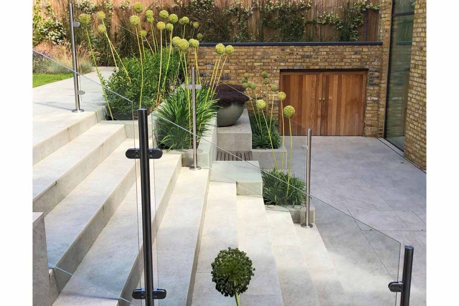 Silver Grey Porcelain Garden Steps descending on to patio, glass panels used as railings with green Alliums planted up the stairs in built in beds.