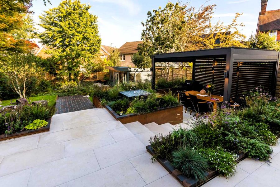 Florence White Porcelain Paving, with steps leading down to black pergola with dining table beneath.