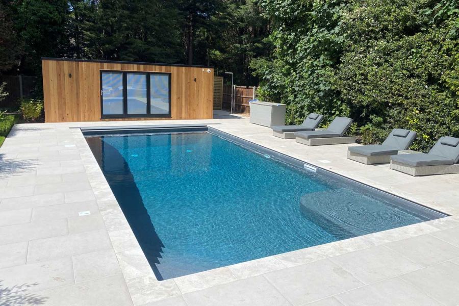 Rattan sun loungers lined up next to large trees, Garden building sits at the end of Florence White Porcelain patio with a swimming pool in the centre.