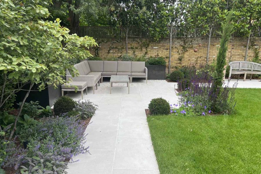 Florence White porcelain path, 2 slabs wide, leads to square paved area in corner of garden with modular sofa and table.