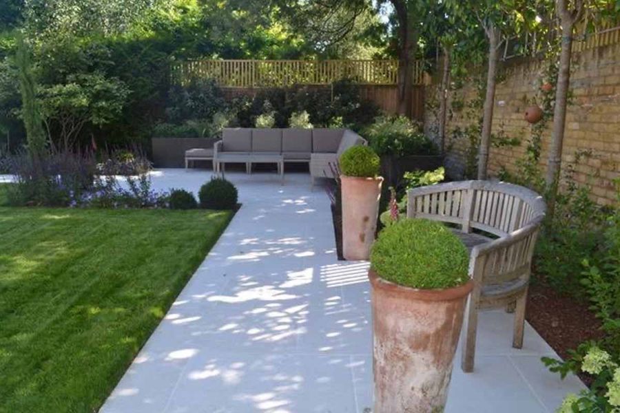Florence White porcelain pathway between wall and lawn connects patio with outdoor sofa to paved area with bench and 2 tall pots.