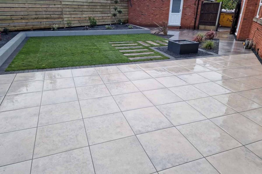 Irregular-shaped garden, half grass, half laid with Jura Grey porcelain paving, edge cut at angle to fit along house wall.