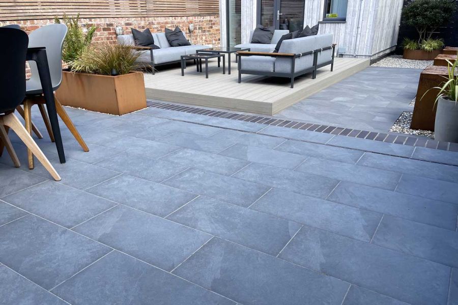 Slab Coke Porcelain patio laid in a stretcher bond pattern next to a raised decking area featuring a light grey lounging furniture set.