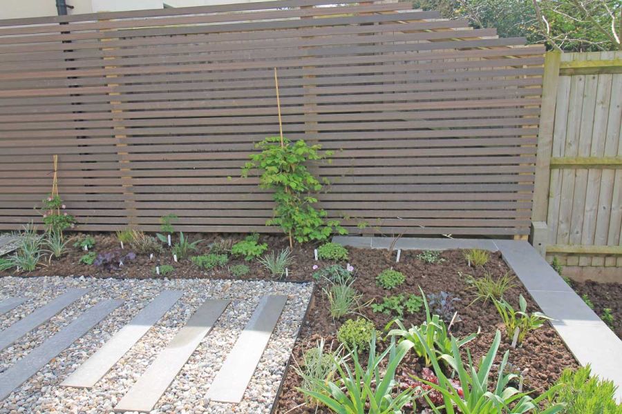 Tall open-slatted screen of Chestnut composite battens attached to wooden fence runs along bed and plank paving set into gravel.