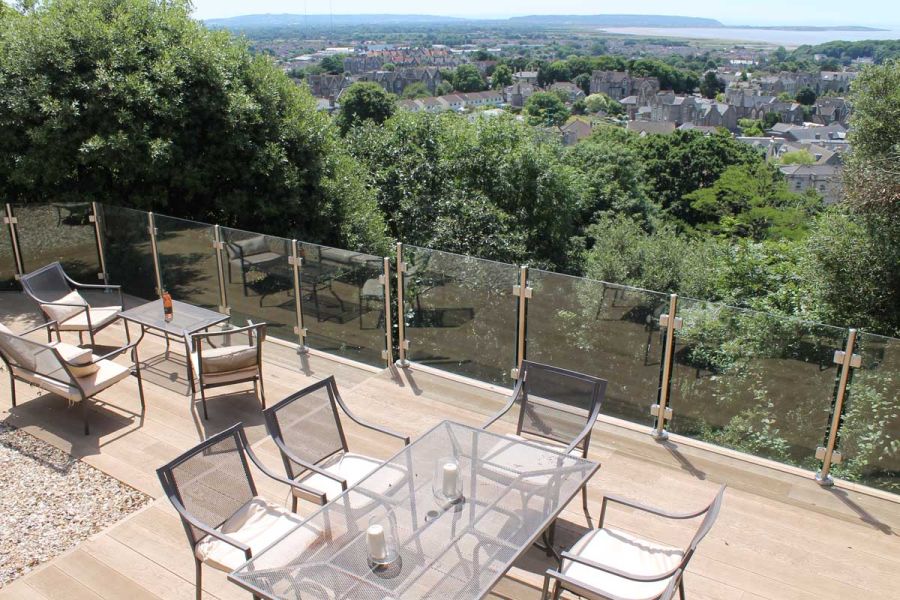 Golden Oak Millboard decking paves roof terrace with glass railings overlooking view to sea. Design by Outerspace Gardens.