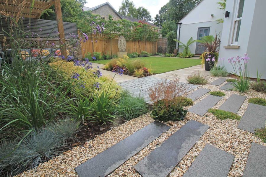 Black granite planks set into buff pea gravel in back garden with lawn and planted beds. Built by Outerspace Creative Landscaping.