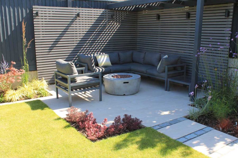 Square paved seating area, Pebble Grey composite batten screening on 2 sides, with path leading away between grass and border. 