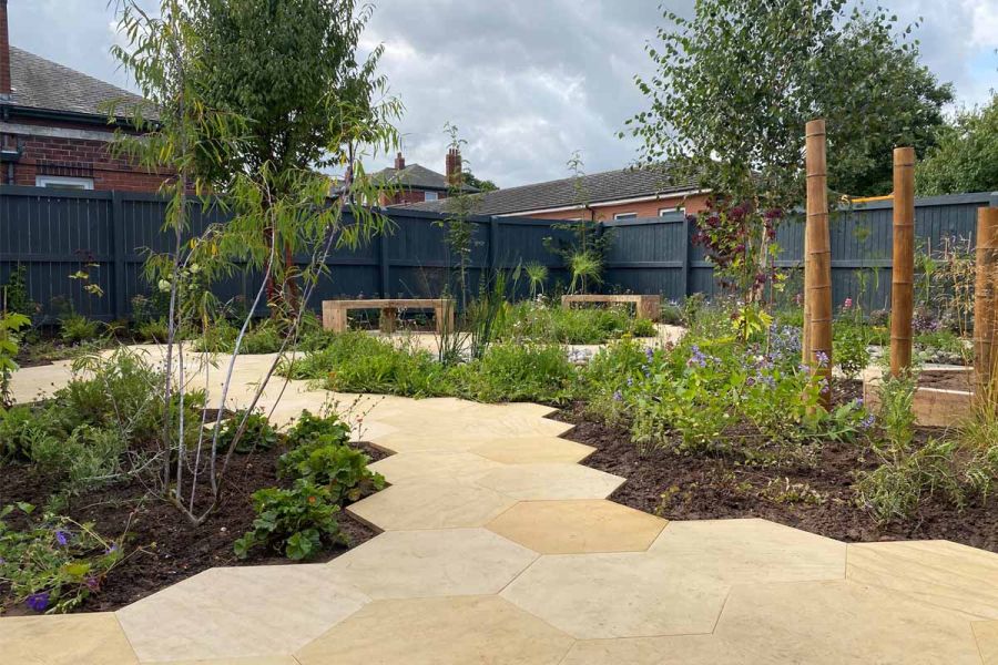 Paths of hexagonal slabs of Dune Smooth Sandstone paving wind past benches and between beds of low planting in grey fenced garden.