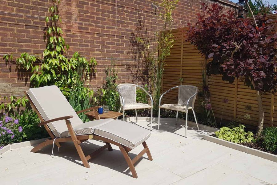 Sun lounger sits on Faro Porcelain Paving next to two rattan armchairs with table in between holding drinks. 