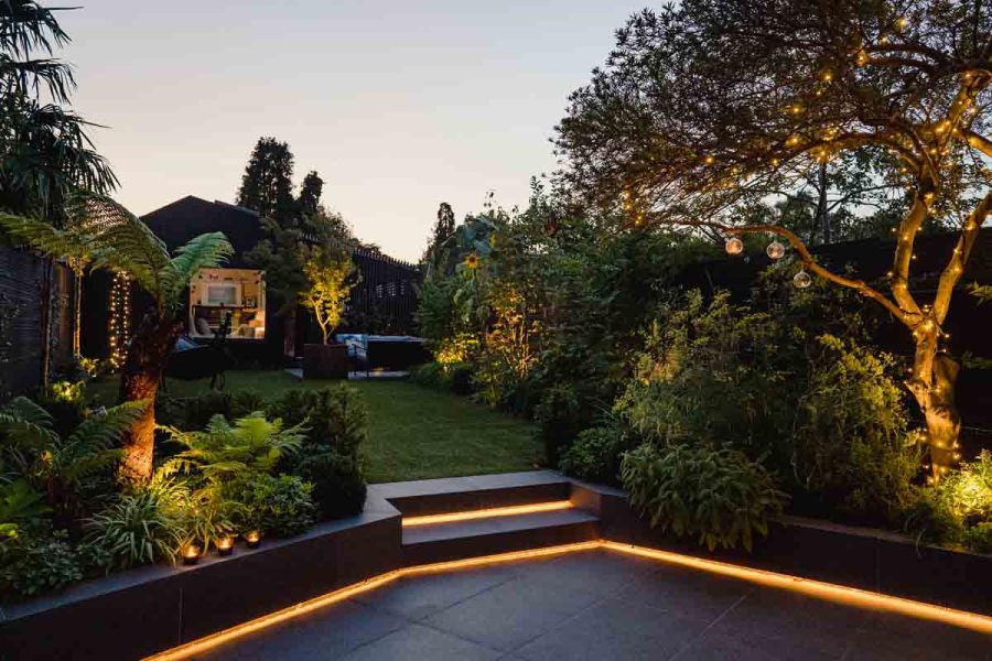 Back garden in twilight with lawn, trees and lush planting. Patio with 2 steps edged with light strips. Design by Jen Berry.