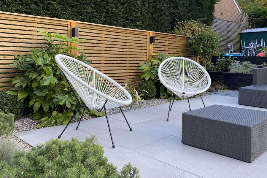 2 wire-strung seats and rattan table sit on Urban Grey porcelain paving edged with planted gravel bed. Design by Creative Gardens.