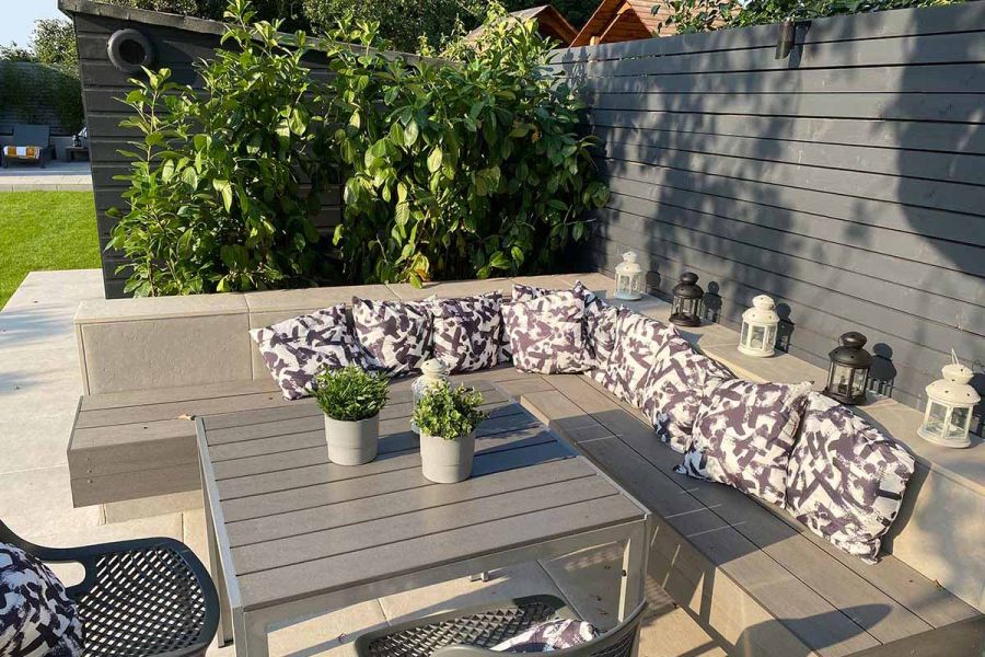 By garden fence, Pebble Grey Brushed Composite Decking tops right-angled cantilevered bench and matching table with potted plants.