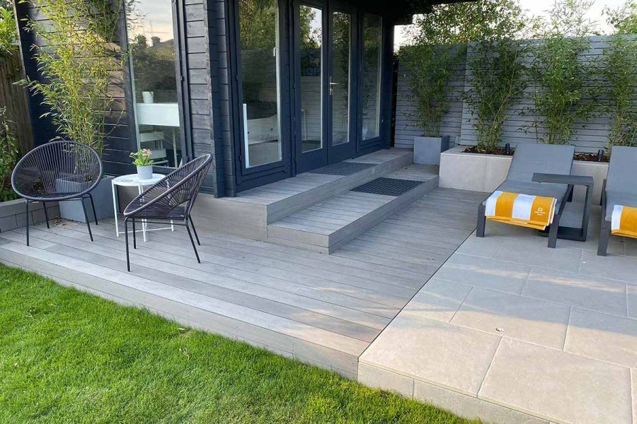 Raised patio of cream porcelain and Pebble Grey Brushed composite decking uk, with 2 steps up to glass-fronted garden room.