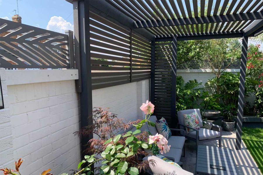 A pergola of spaced Pebble Grey Composite Battens covers paved area with outdoor lounge furniture, in corner of sunny garden.
