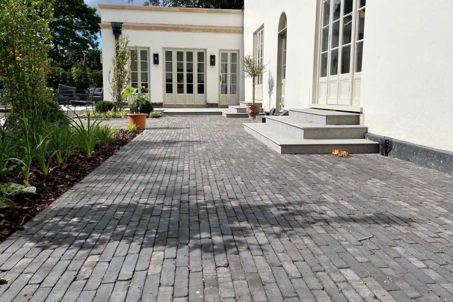 Large patio, built by Landscapes by Design of grey Lucca brick paving, stretches across back of white House with french doors.