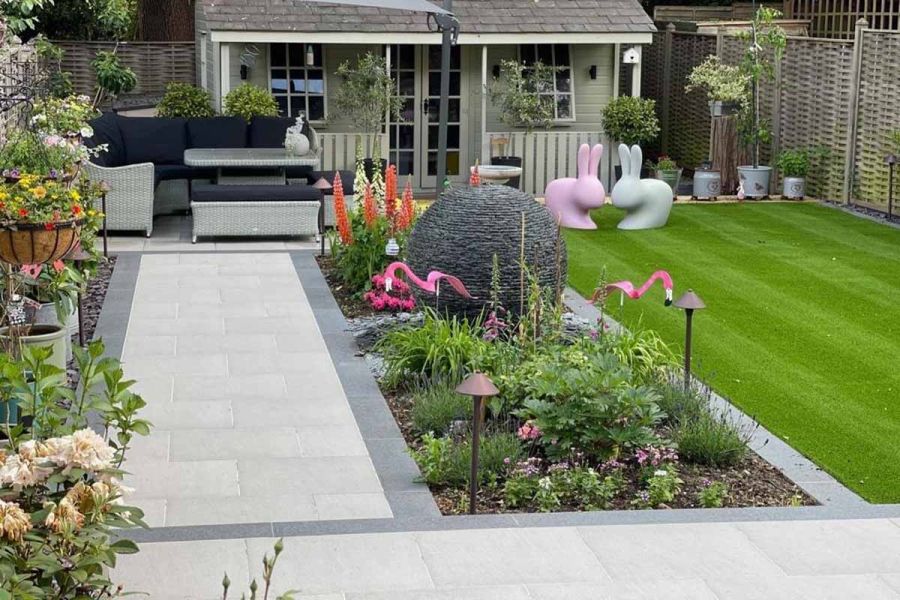 Flower bed separates lawn from path leading to seating area paved in Urban Grey porcelain tiles, garden room behind.