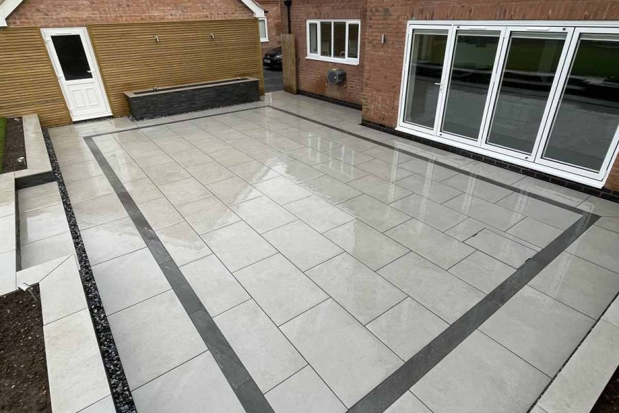 Wet oblong grey patio with inset black outline of Platinum Grey porcelain planks outside french doors of red brick modern house.