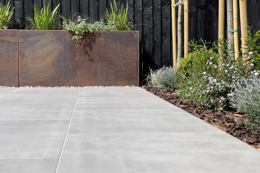 Polished Concrete porcelain 600x600 slabs laid in stretcher bond pattern with a planted flower bed on one side and Corten Steel Planters.