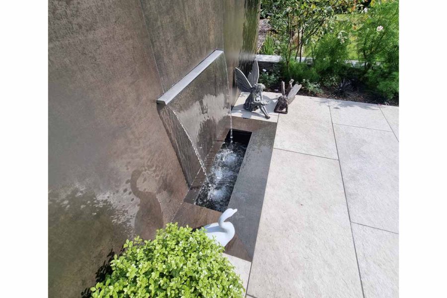 Water feature wall faced in Steel Dark porcelain cladding, with wide spout pouring water into rectangular pond beneath. 