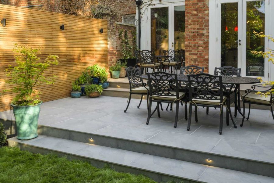 French doors overlook patio of Florence storm porcelain outdoor tiles uk, with 2 steps down to lawn. Design by Jaki Grosvenor. 