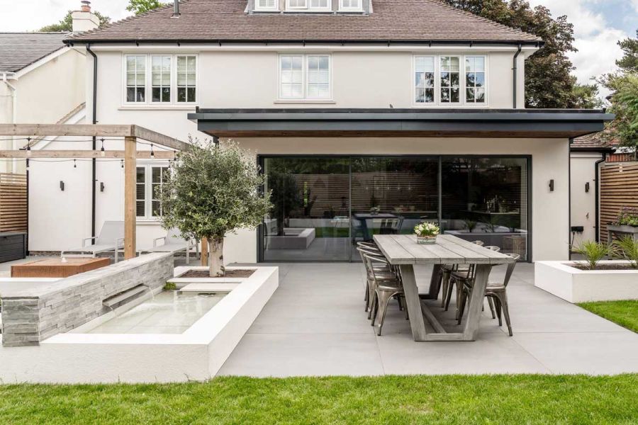 Light grey patio with long table next to raised rectangular pond laid with Comblanchien porcelain tiles. Built by Gadsden Gardens.