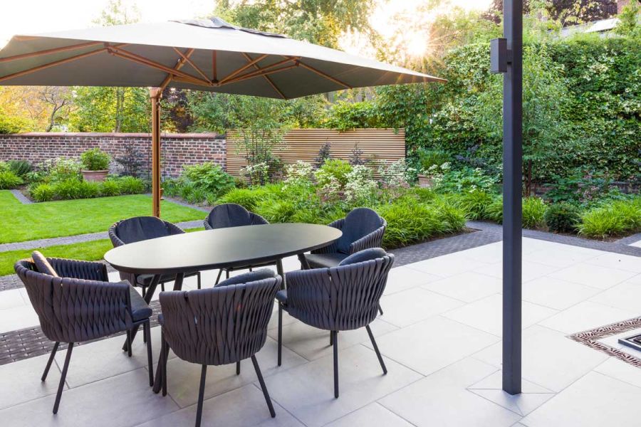 Parasol over table and 6 chairs on Florence Grey porcelain paving with clay paver edging. Rectilinear planted beds next to lawn.