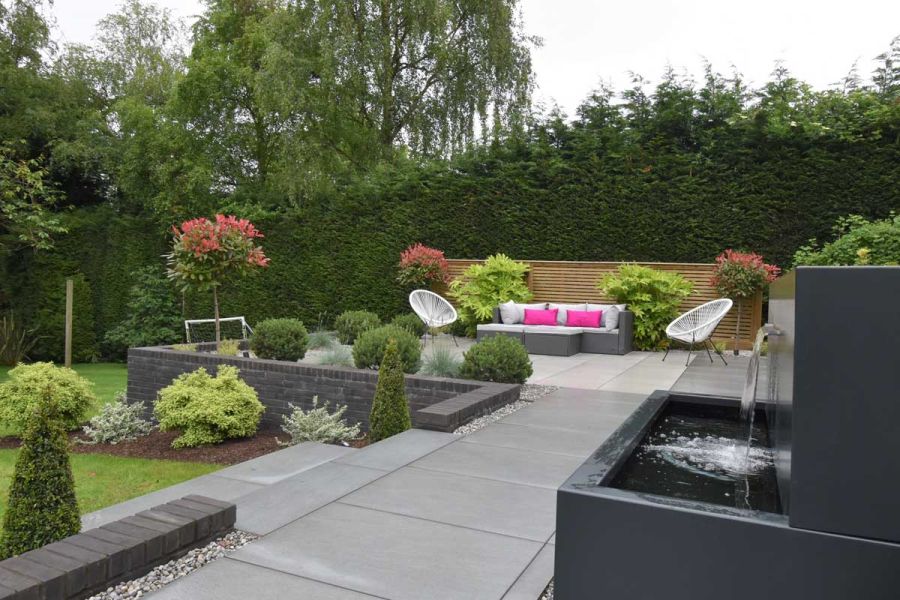 Water falls into trough from spout by path of Trendy Black porcelain paving leading to seating area and shrubs. Steps down to lawn.