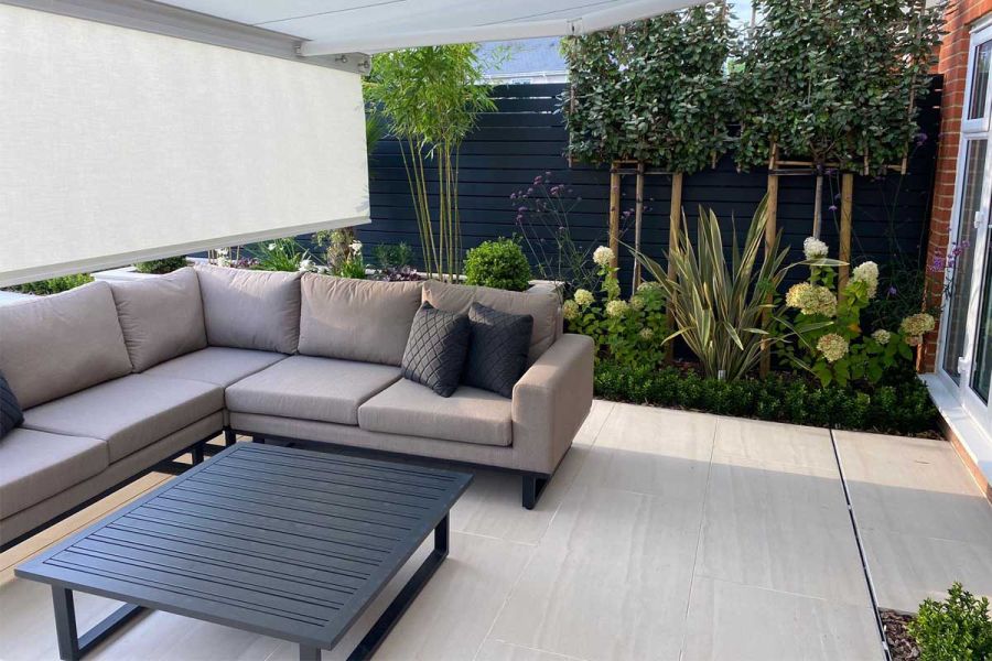 Grey modular sofa and coffee table on Faro Porcelain patio tiles with slot drain, in front of bed of hydrangeas and boxhead trees.