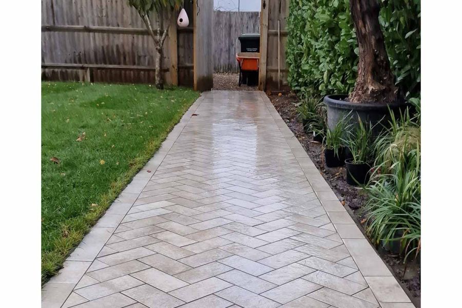 Open wooden gate in fence shows black wheely bin at end of path of Cream porcelain patio tiles laid herringbone in lawned garden