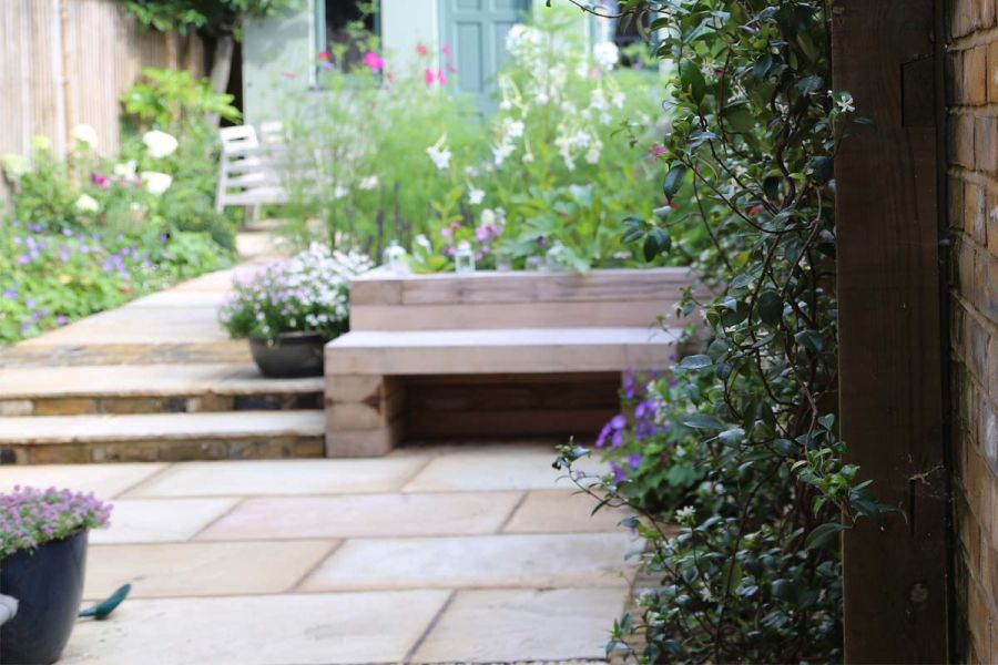 Mint Indian sandstone paving with wooden bench to one side of 2 steps up to path leading to back of house between colourful planting.