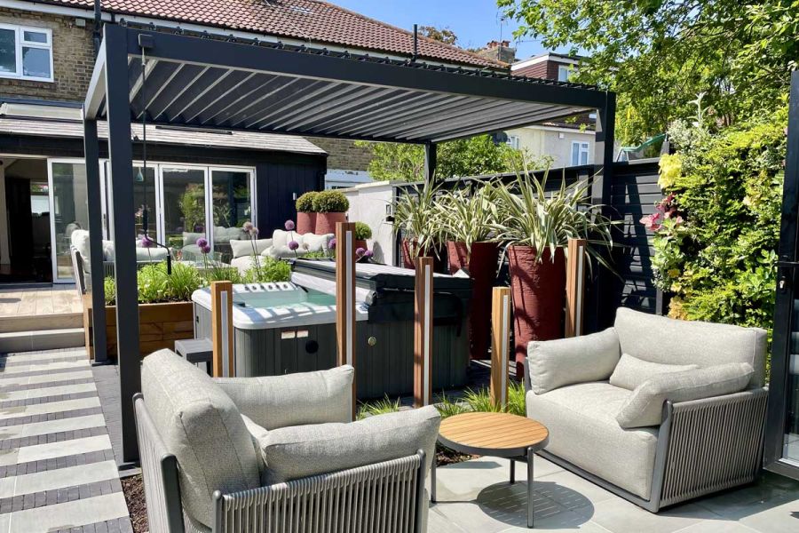 Potted plants sit on hot tub on brick paving under Proteus grey aluminium pergola, next to 2 armchairs and striped path.