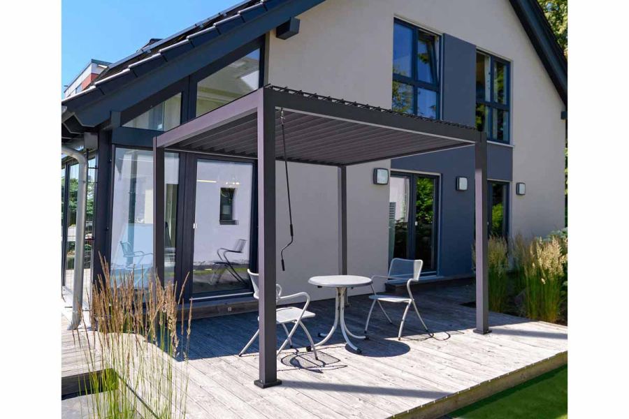 Proteus Grey Aluminium Pergola on decking next to house; table and 2 chairs beneath; crank handle for opening louvres in place.