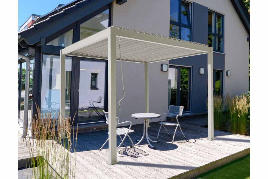 A Proteus White Metal Pergola with louvred roof stands on decking next to glass doors into house, covering a table and 2 chairs.