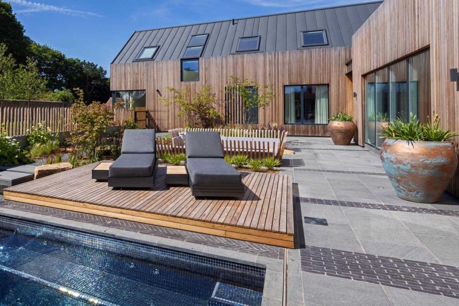 Modern wooden house on 2 sides of large patio paved with slabs and white stripes of Amersham clay pavers next to raised decking.