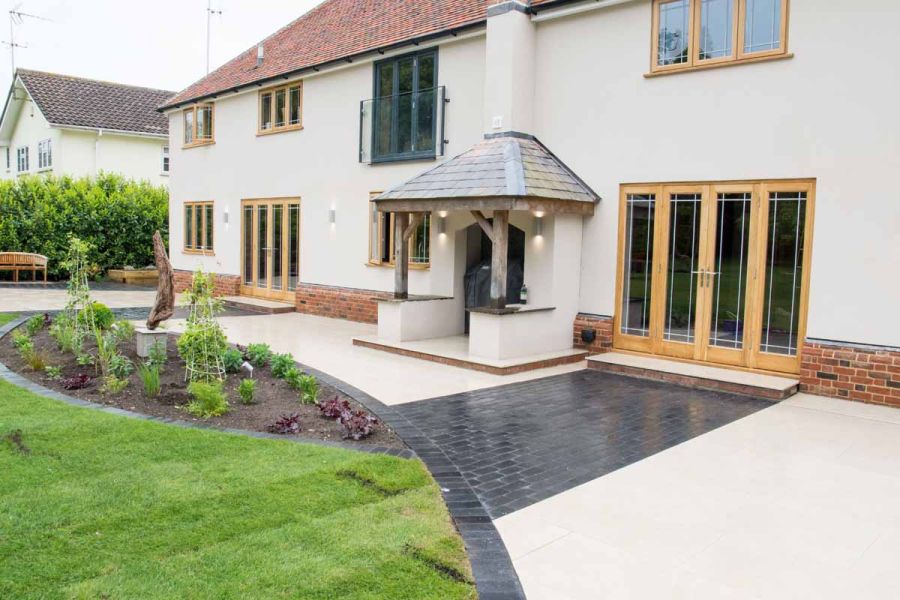 Cream coloured house with wooden porch entrance uses golden stone porcelain paving with black limestone setts in curved design.
