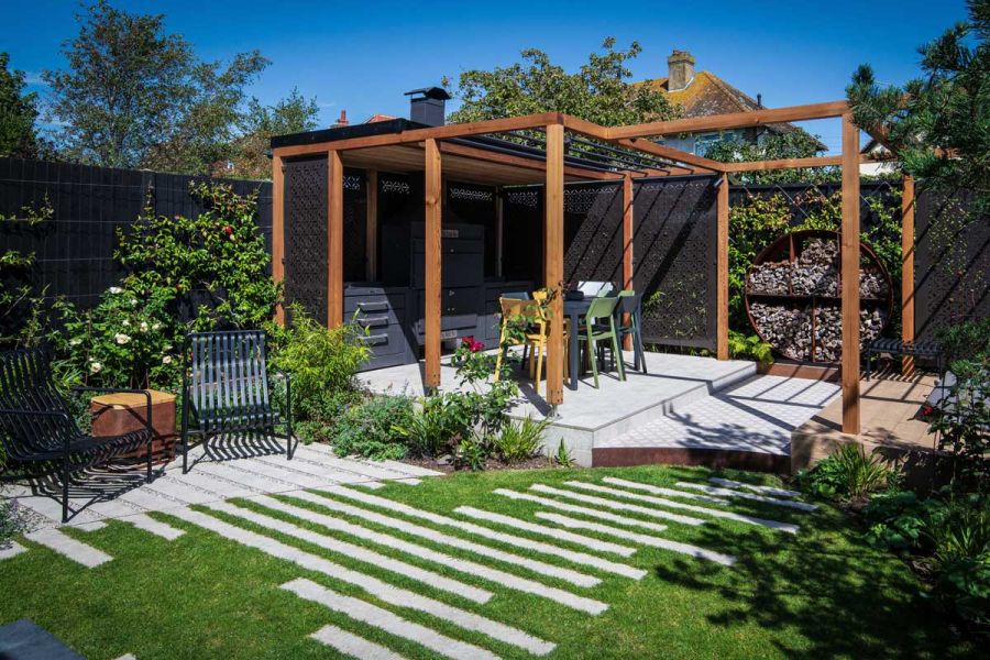Corner of fenced garden. Light Grey porcelain plank paving set into curved lawn, next to raised patio covered by wooden pergola.