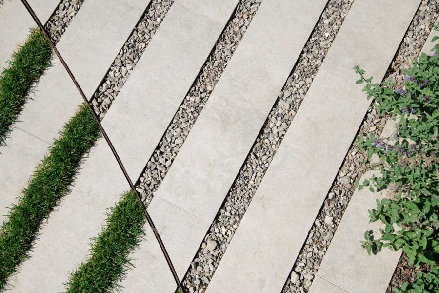 Light Grey porcelain plank paving bisected by curved metal filet, wide gravel joints on 1 side, grass on other. Design by R Goozee.