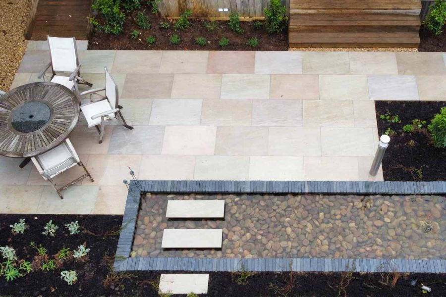 Birds eye view of Raj Green Porcelain Paving next to pebble lined pond clad with black bricks, garden furniture to the left.
