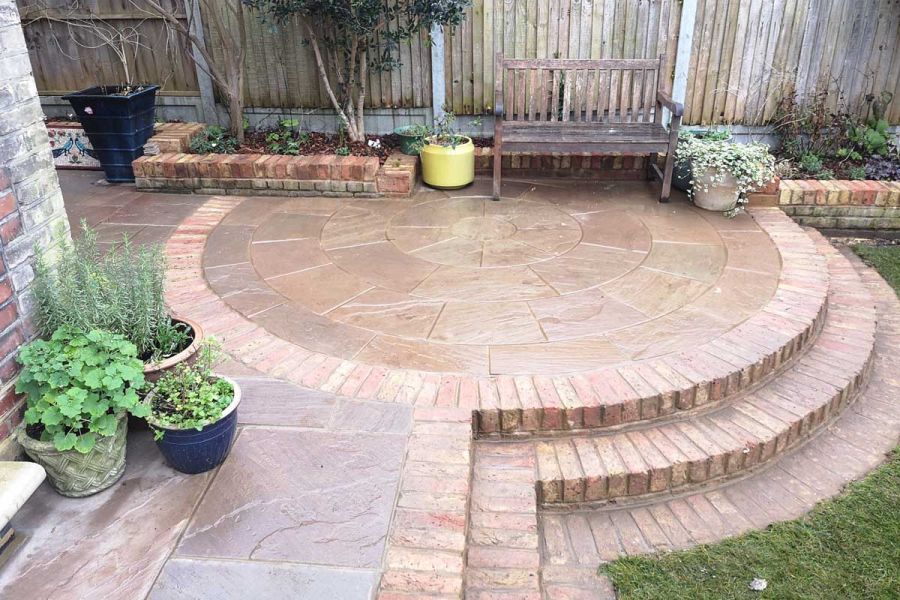 2 curved brick steps up to Raj Green Sandstone paving circle outlined with same bricks in patio with pots, bench and raised bed.