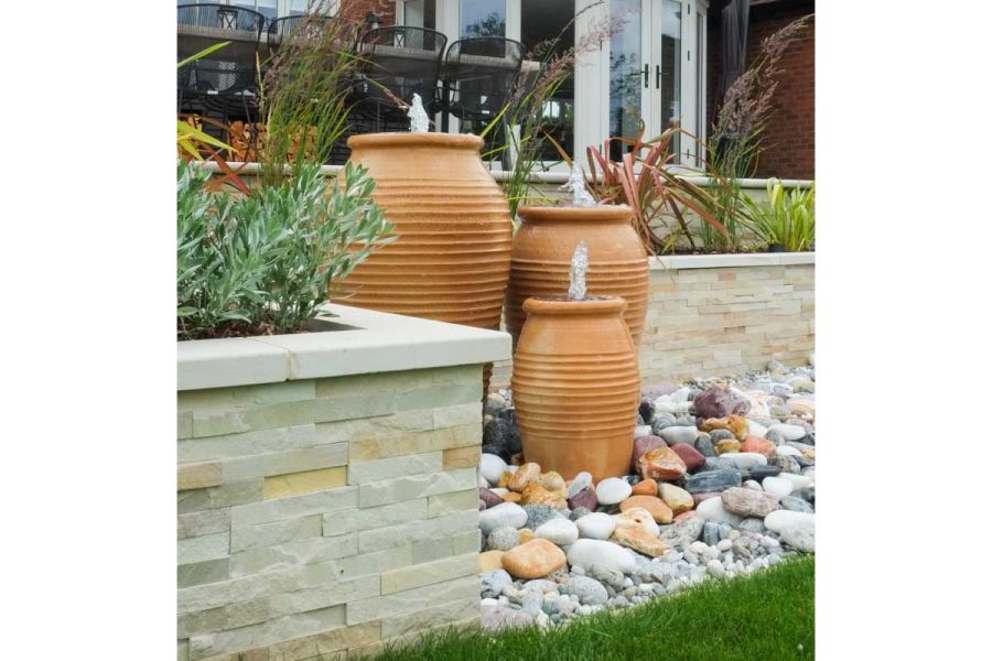 Water feature of 3 terracotta pots sits on large pebbles in right-angled corner of raised bed faced with Mint sandstone cladding.