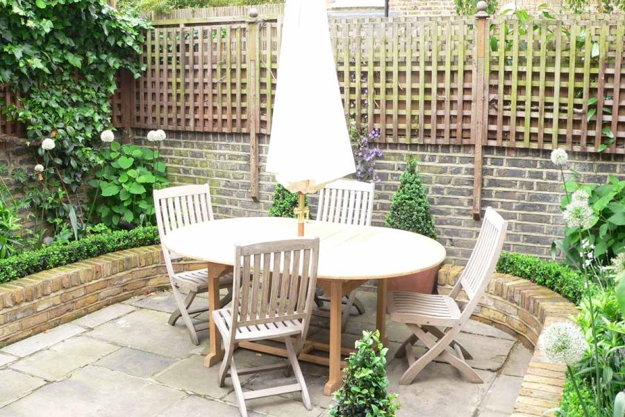 Oval wooden table and 4 chairs on reclaimed Yorkstone patio bordered by low brick wall with small clipped hedge behind.