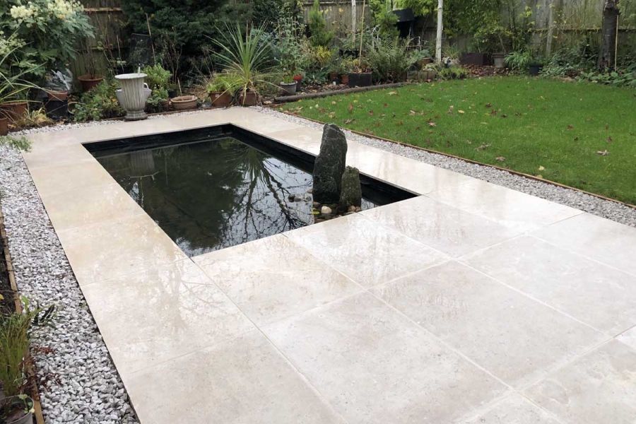 2 standing stones in rectangular pond set in Egyptian beige limestone paving, edged with gravel next to lawn. 