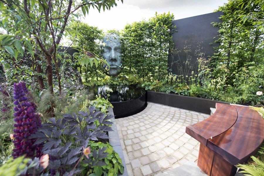 Wall faced in Steel Dark porcelain cladding interrupts hedge in show garden with round raised pond, bench and sculpture.