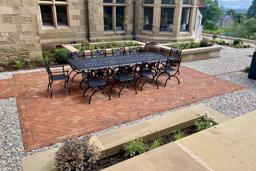 Dining set on oblong area of Romsey Antique Belgian bricks laid herringbone pattern next to stone raised beds of old house.