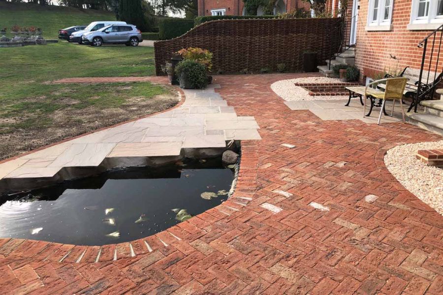 Patio of riven stone and Romsey Antique clay pavers, with pond and table and chair on edge of grassed area with parked cars.