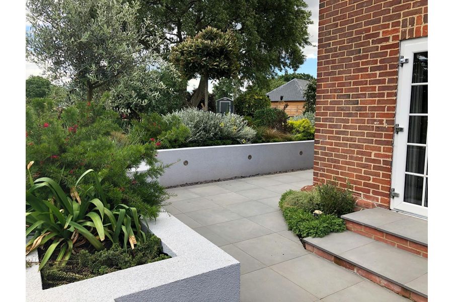 Steel Grey porcelain patio wrapping around the side of a red brick house with a rendered retaining wall to house raised planted beds.
