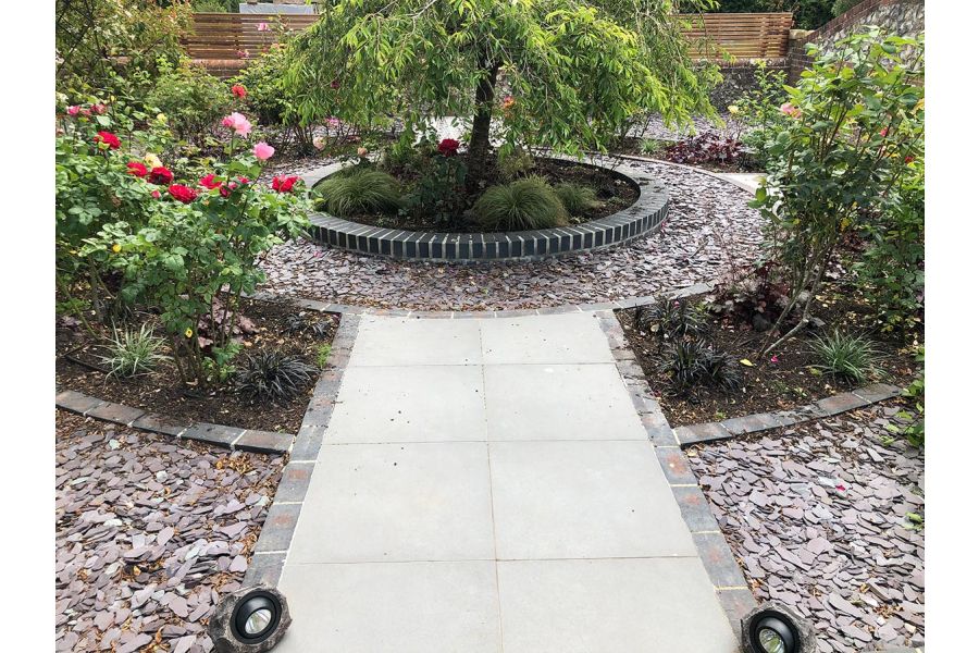 Steel Grey porcelain path leading to a circular brick bed with a tree as the centre piece and surrounding rings of slate chipping and flower beds.