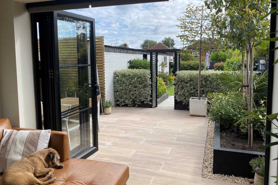 Dog rests on brown leather couch inside, french doors wide open looking onto Rovere Porcelain paving patio.