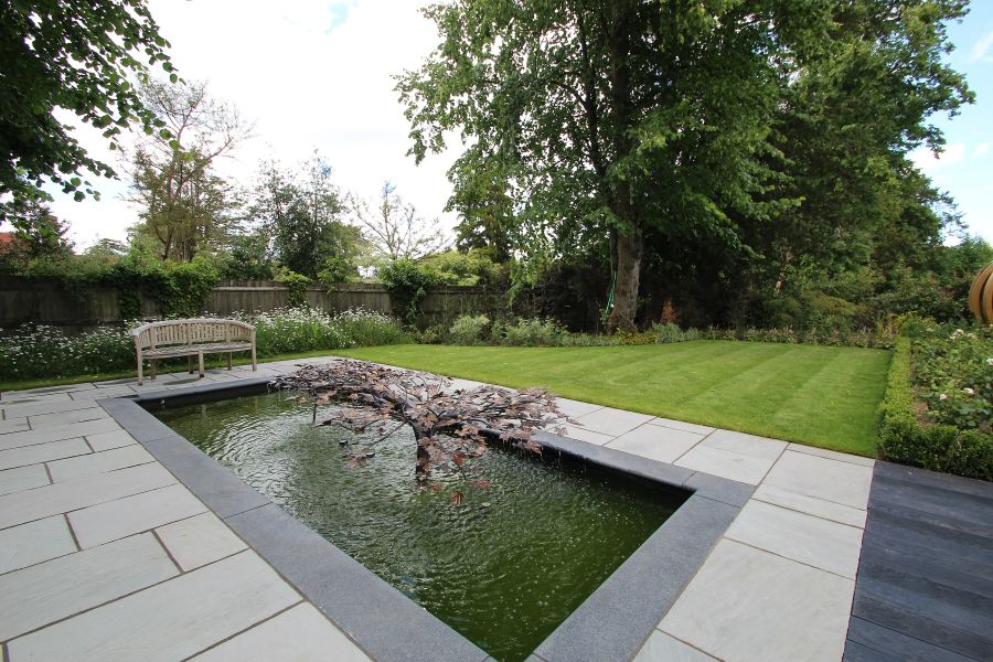 Tree water feature in rectangular pond with Kandla Grey tumbled sandstone surround, next to lawn with borders edged with low hedge.