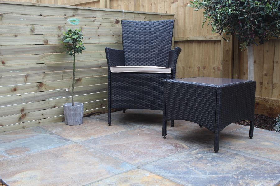 Single rattan armchair with cream cushion sits next to matching glass topped table, on top of rustic slate paving.